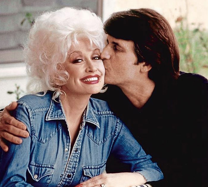 Dolly Parton's sister smiling in denim jacket while receiving a kiss, raising alarm amid singer health struggles.