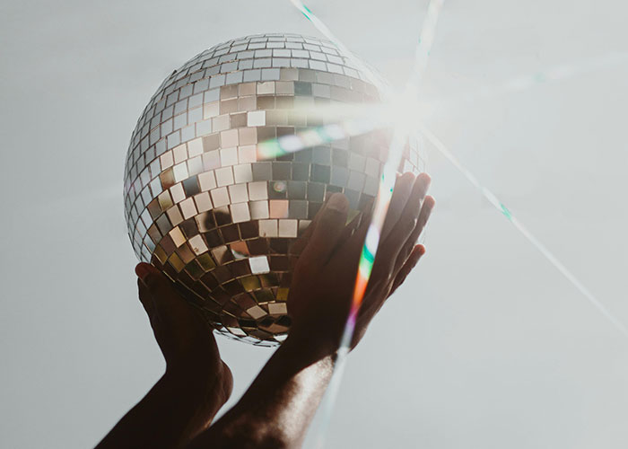 Hands holding a reflective disco ball with bright sunlight creating lens flare and colorful light streaks.