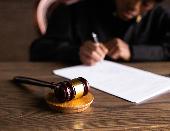 Gavel lying on wooden table in courtroom setting with blurred person writing legal document in background.