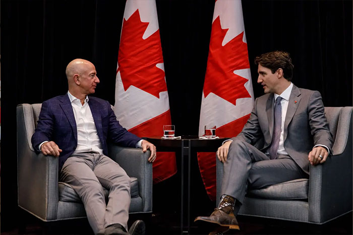 Justin Trudeau in a formal suit sitting with Jeff Bezos against a backdrop of Canadian flags during a discussion.
