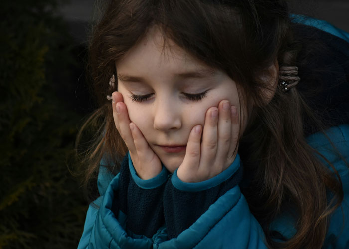 Young girl with eyes closed and hands on cheeks, expressing a thoughtful mood related to plastic surgeons and surgeries.