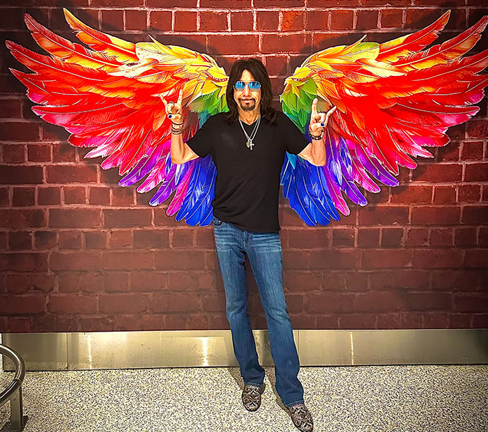 Man wearing sunglasses and black shirt posing with colorful painted wings on a brick wall, related to Gene Simmons and Paul Stanley.