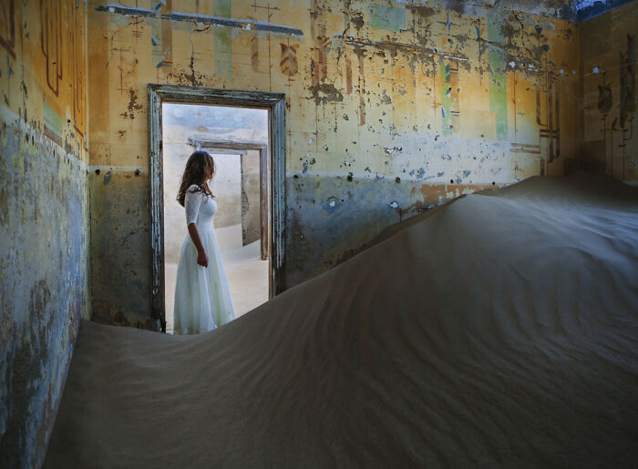 Woman in white dress inside abandoned home filled with desert sand in surreal sand castles photo series
