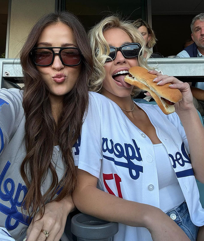 Two women wearing sunglasses and Dodgers jerseys at a game, one holding a hotdog, reflecting pro-modesty fashion debate.