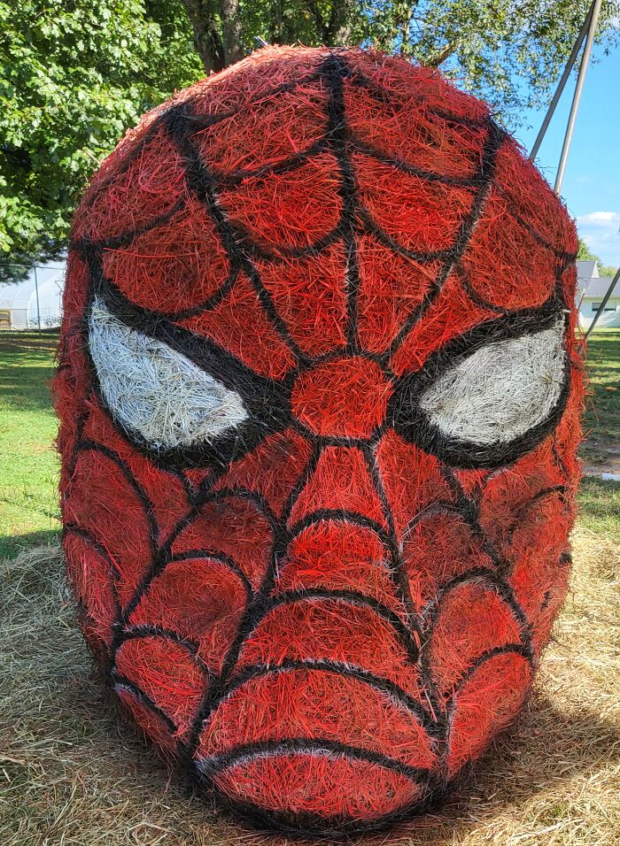 Large Halloween hay bale art painted as Spider-Man face with red, black, and white colors outdoors on grass under trees.