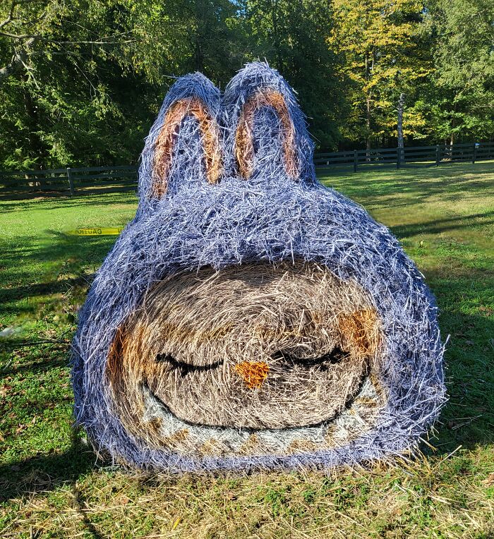 Hay bale art painted as a Halloween character with closed eyes and bunny ears in an outdoor grassy area.