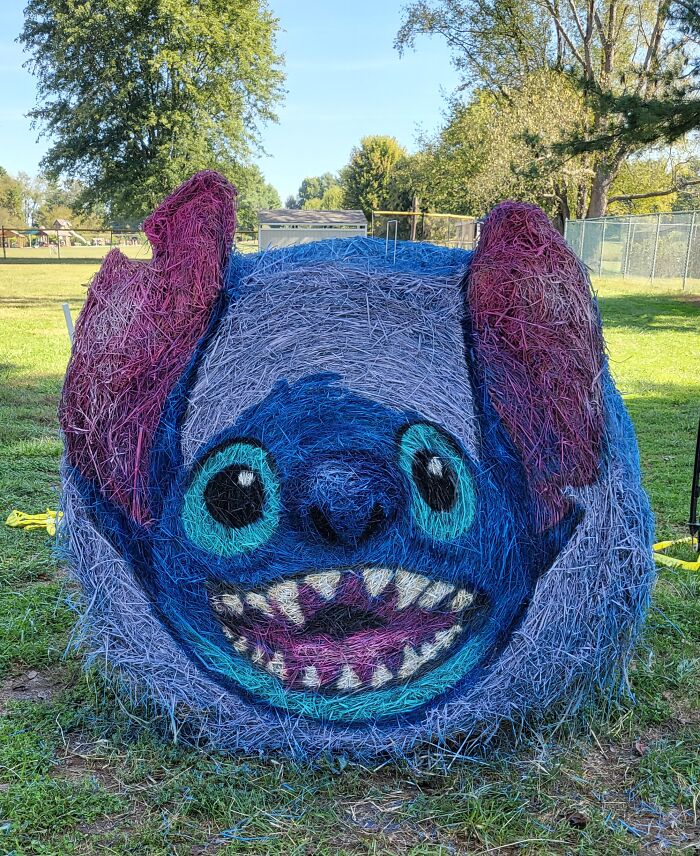 Colorful Halloween hay bale art painted with a playful cartoon face featuring large eyes and sharp teeth outdoors on grass.