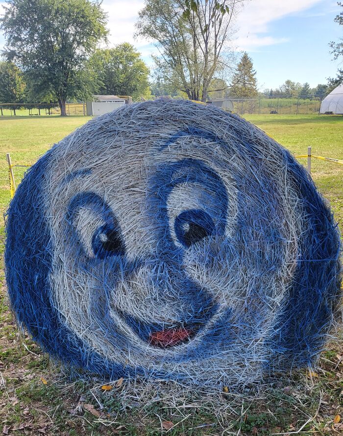 Halloween hay bale art featuring a friendly ghost face painted in blue and white on a round hay bale outdoors.