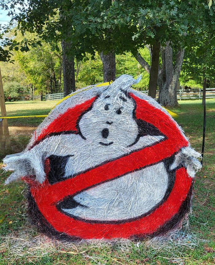 Halloween hay bale art painted with a ghost and red no symbol, displayed outdoors among trees and grass.