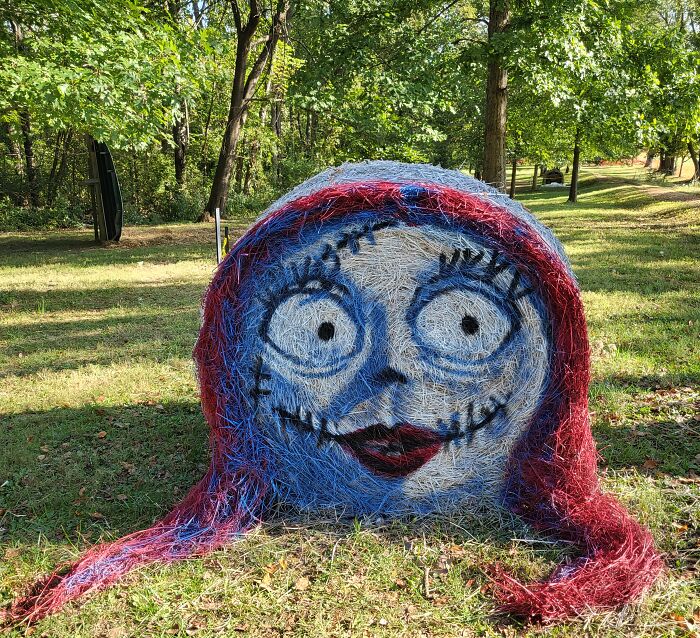 Halloween hay bale art painted with a cartoon face featuring big eyes and stitched smile in an outdoor grassy area.