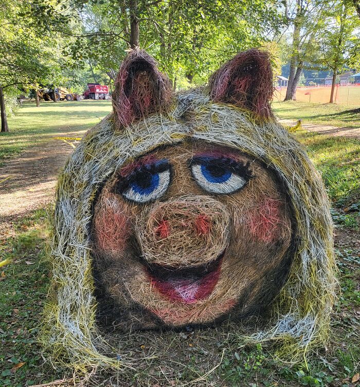 Pig face Halloween hay bale art with blue eyes and blonde hair displayed outdoors in a park setting for kids and families.