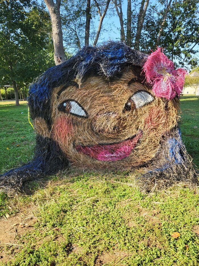 Halloween hay bale art featuring a smiling face with painted eyes, pink lips, and a pink bow in an outdoor park setting.