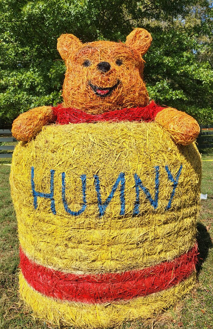 Hay bale art of a bear in a red shirt inside a yellow "Hunny" pot, a creative Halloween hay bale decoration outdoors.