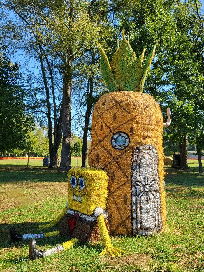 Halloween hay bale art depicting a pineapple house and cartoon character in a sunny park setting with trees and grass.