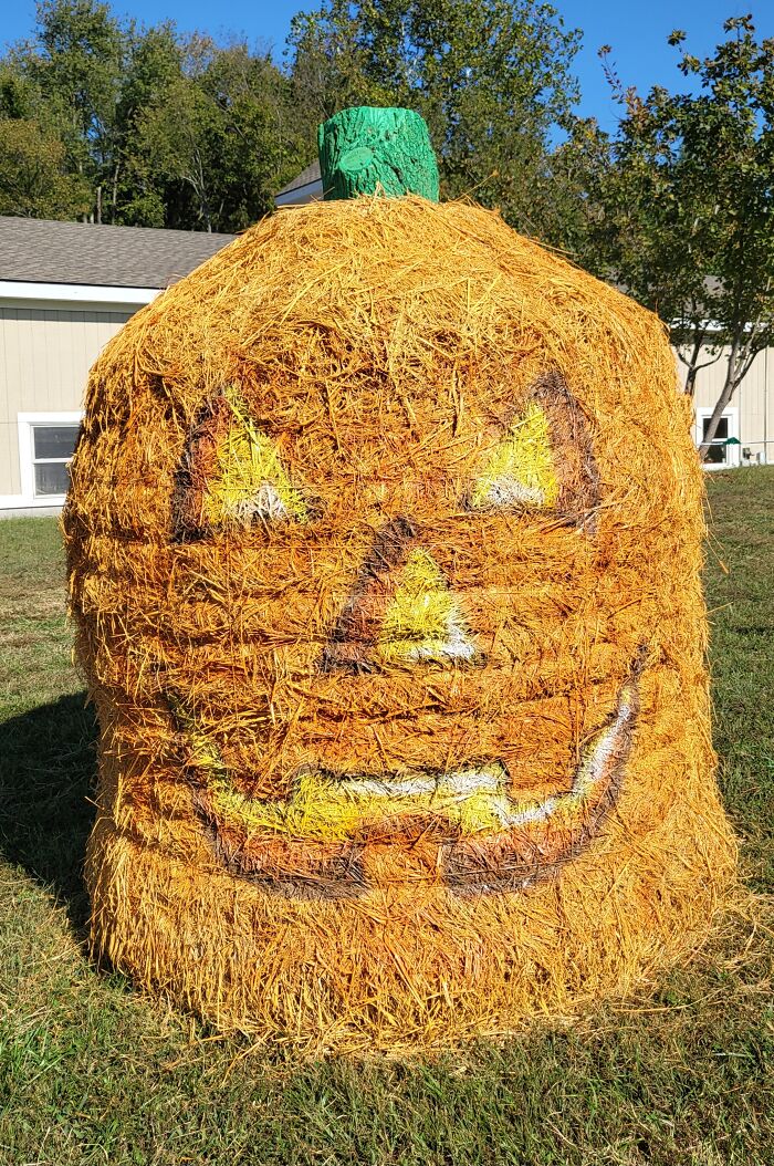 Large Halloween hay bale art painted as a jack-o'-lantern with a green stem outdoors on grass.
