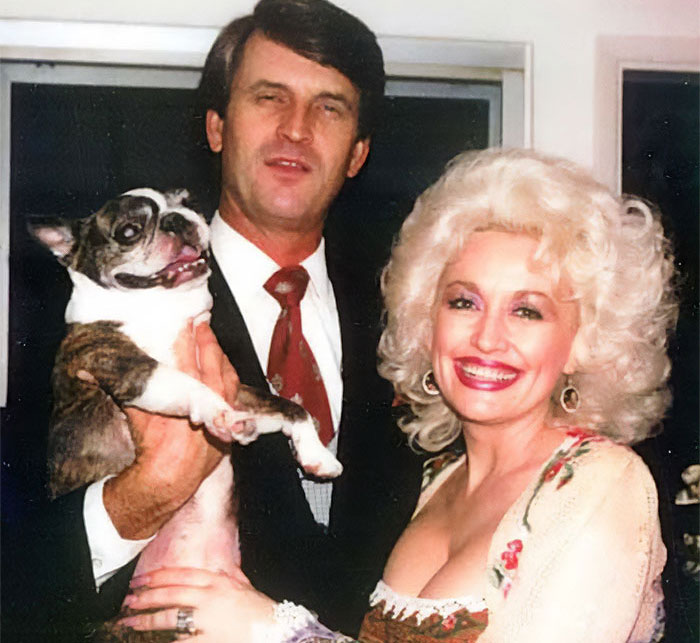 Dolly Parton smiling beside a man holding a dog, highlighting sister raising alarm amid singer health struggles.