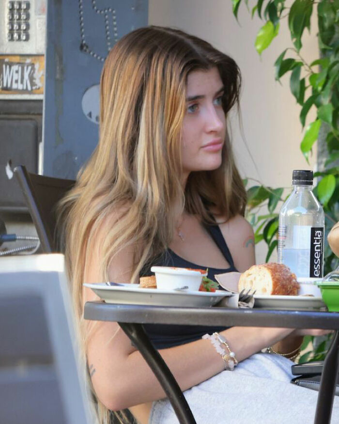Charlie Sheen's daughter sitting at an outdoor table, showing natural look before and after plastic surgeries.