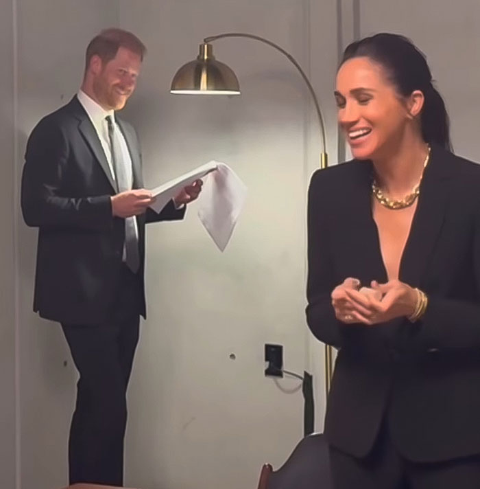 Meghan Markle and Prince Harry dressed formally indoors, with Meghan smiling and Prince Harry holding papers.