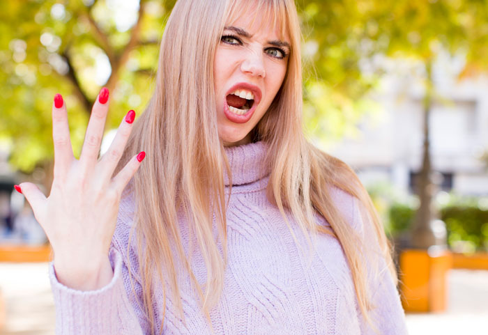 Angry woman with long blonde hair and red nails showing frustration outdoors, representing cruel person emotions.