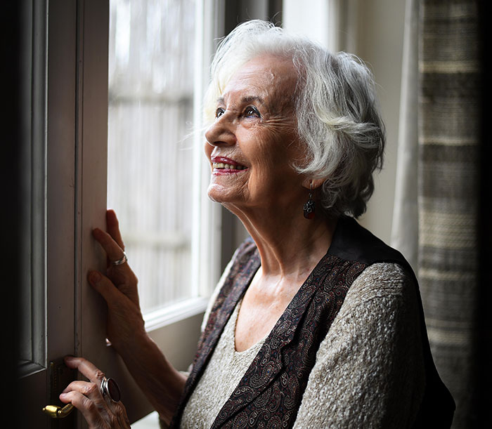 Holocaust survivor Ruth Posner smiling gently while standing near a window, reflecting on her life and memories.