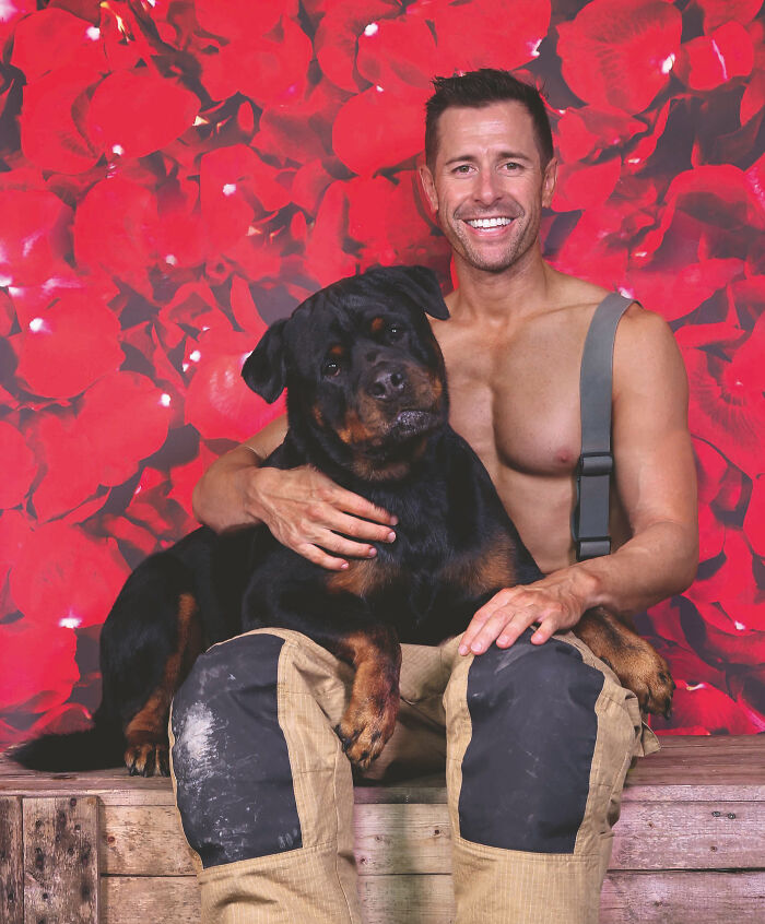 Firefighter in suspenders smiling and sitting with a large black and brown dog against a red floral backdrop.