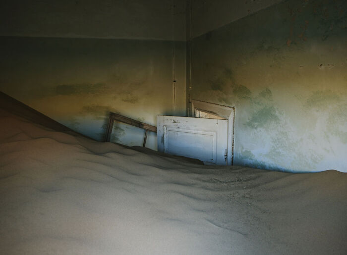 Desert sand fills an abandoned home interior, covering the floor and partially burying doors in a surreal sand castle scene.