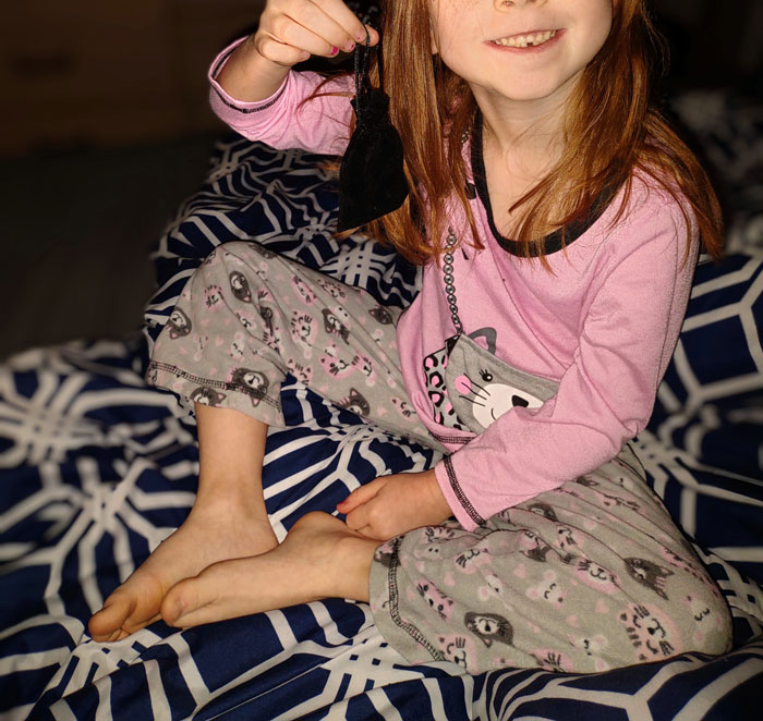 Young girl in pajamas sitting on a patterned blanket, smiling while holding a small black pouch, symbolizing resilience from narcissistic mothers.