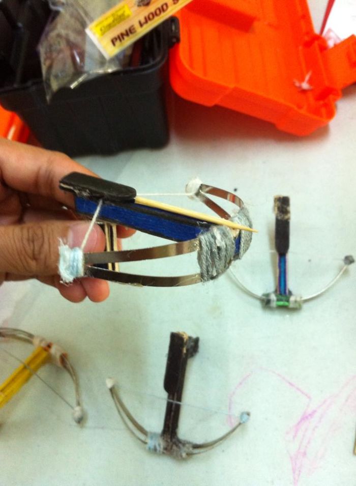 Hand holding a small homemade crossbow invention, with other DIY mini crossbows on a white surface nearby.
