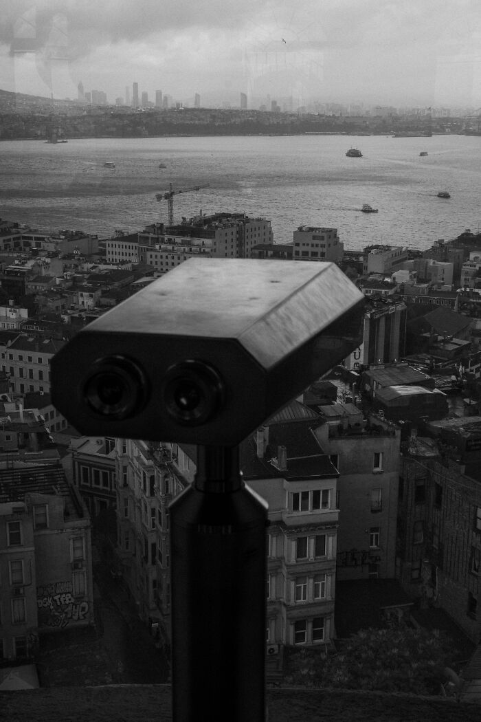Black and white street photography shot featuring a cityscape with a binocular viewer overlooking buildings and water.