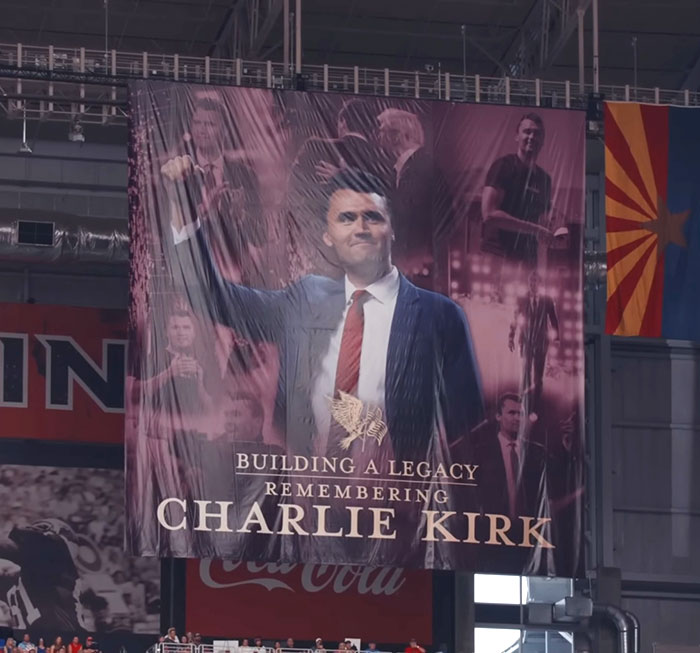Large banner of Charlie Kirk in a suit and red tie inside a stadium, related to a controversial homework assignment.