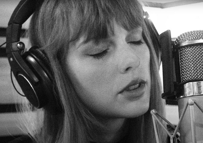 Black and white image of Taylor Swift wearing headphones and singing into a microphone in a recording studio.