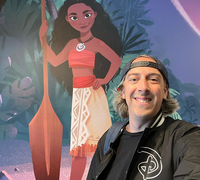 Man wearing a backward cap and Disney jacket smiling in front of Moana character poster amid backlash over heterosexual breeding propaganda. Man wearing a backward cap and Disney jacket smiling in front of Moana character poster amid backlash over heterosexual breeding propaganda.