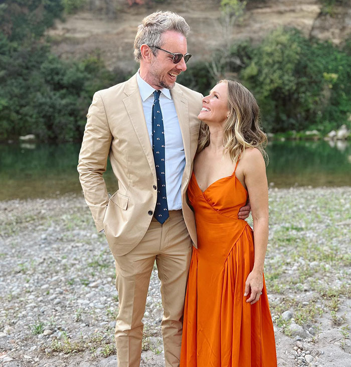 Kristen Bell smiling in an orange dress standing outdoors with a man in a tan suit amid expert backlash and triggering post debates.
