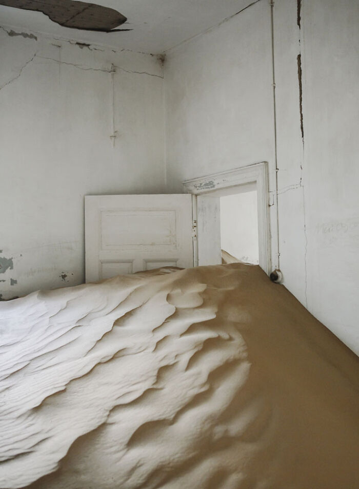 Desert sand filling an abandoned home interior, with a partially open door and cracked white walls.