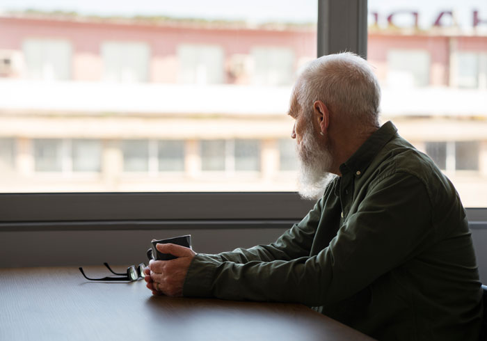 Elderly man with a white beard sitting by a window, reflecting quietly, symbolizing endurance of narcissistic mothers.