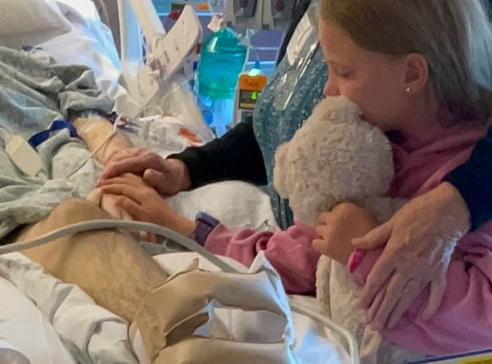 Florida dad-to-be in hospital bed holding hands with child who hugs a teddy bear during emotional visit. Florida dad-to-be in hospital bed holding hands with child who hugs a teddy bear during emotional visit.