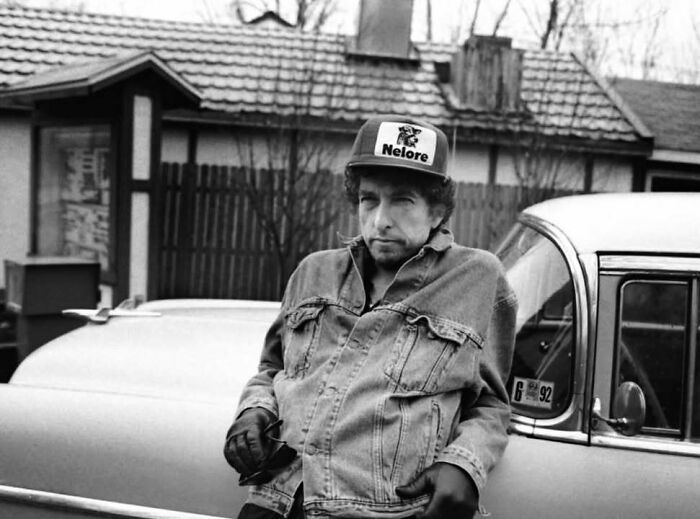 Black and white photo of a man in a denim jacket and cap leaning on a car, representing movie and music industries.