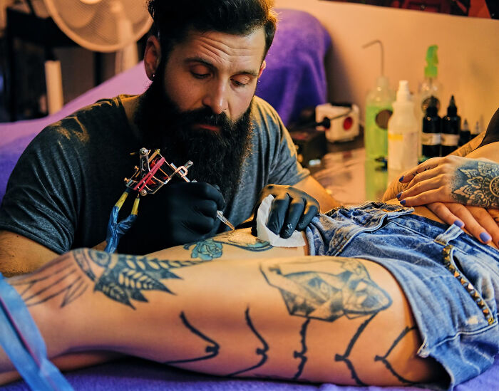 Tattoo artist focused on inking a detailed design on a person's leg in a colorful tattoo studio setting.