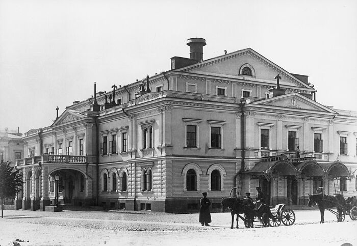 Historic black and white photo showing a grand building and horse-drawn carriages in a world capital before modern times.