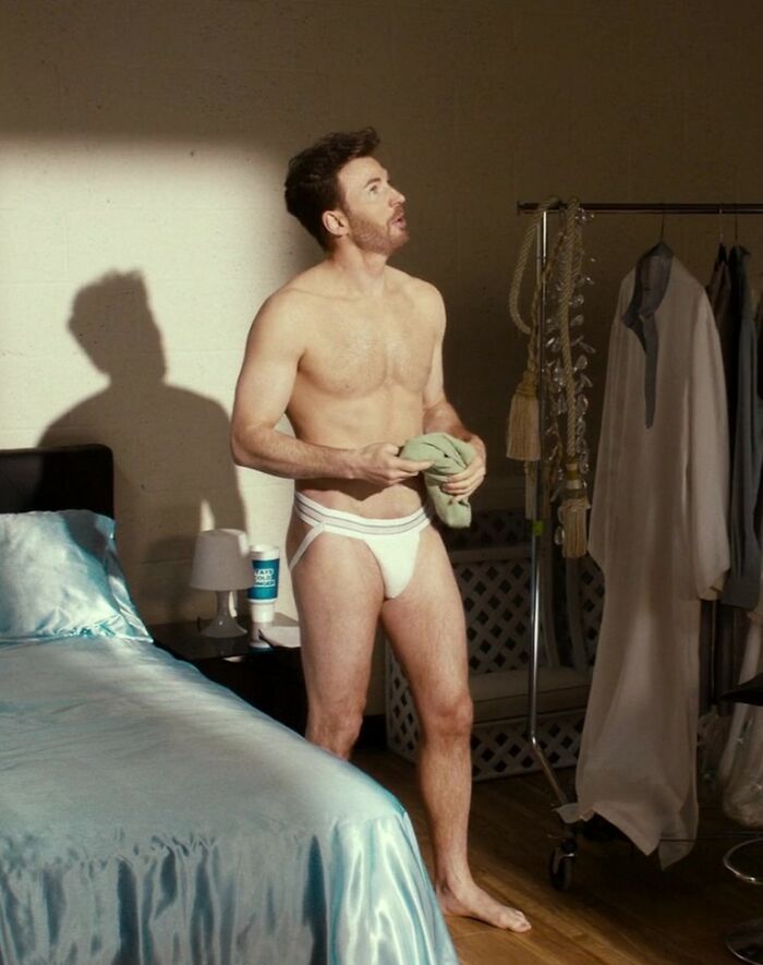 Chris Evans standing shirtless in a white jock strap in a bedroom, holding a green cloth and looking up.