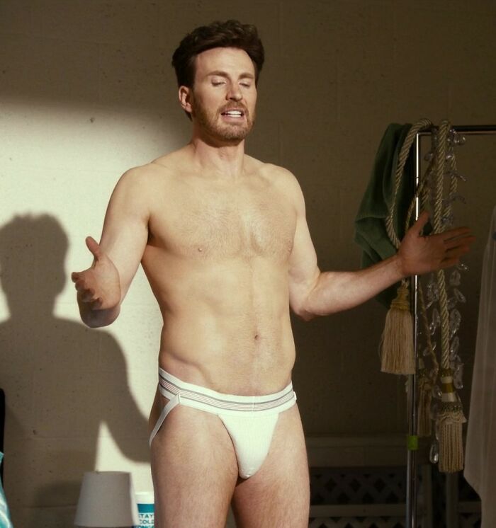 Chris Evans posing shirtless wearing a white jock strap with arms open in an indoor setting.