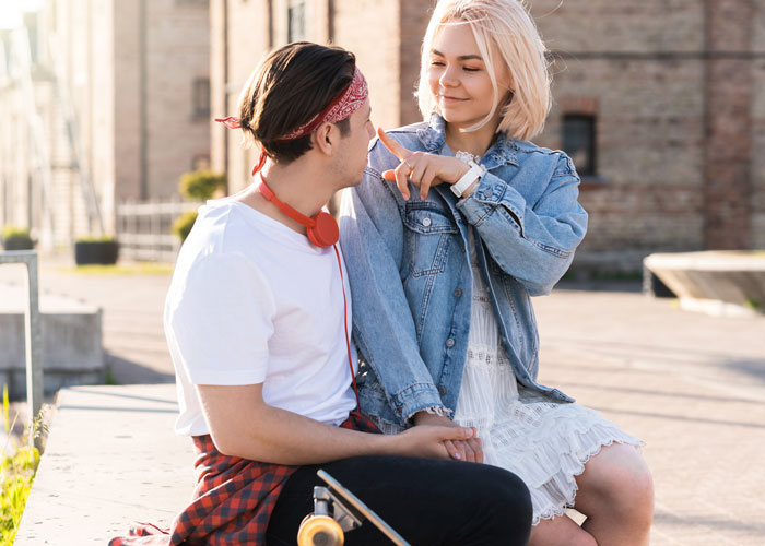 Young couple sitting outdoors, man holding skateboard, woman gently touching his nose, showing true colors after breakup.
