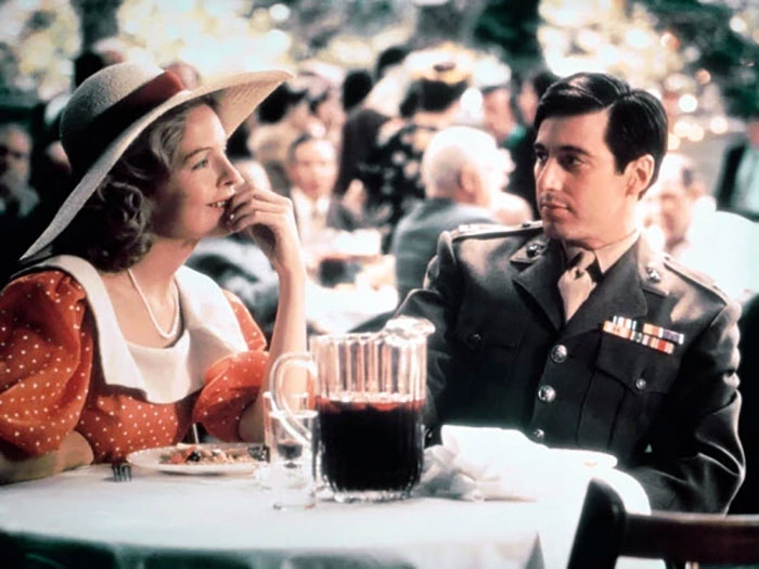 Scene with Diane Keaton and a man in military uniform seated at an outdoor table, vintage setting reflecting a past era.