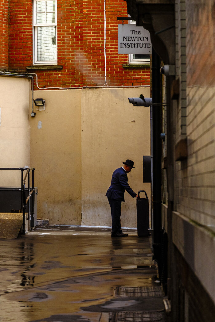 Man in a suit and hat interacts with a trash bin in a narrow urban alley, showcasing stunning street photography moments.