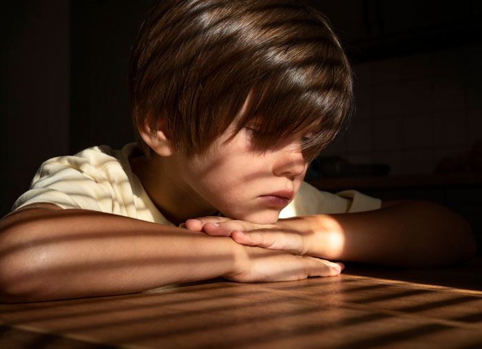 Young boy sitting at table with arms crossed, looking down, illustrating feelings of cruelty and emotional hurt.