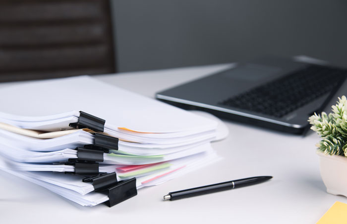 Stack of documents with binder clips, pen, laptop, and plant on a desk, related to plastic surgeons and unnecessary surgeries.
