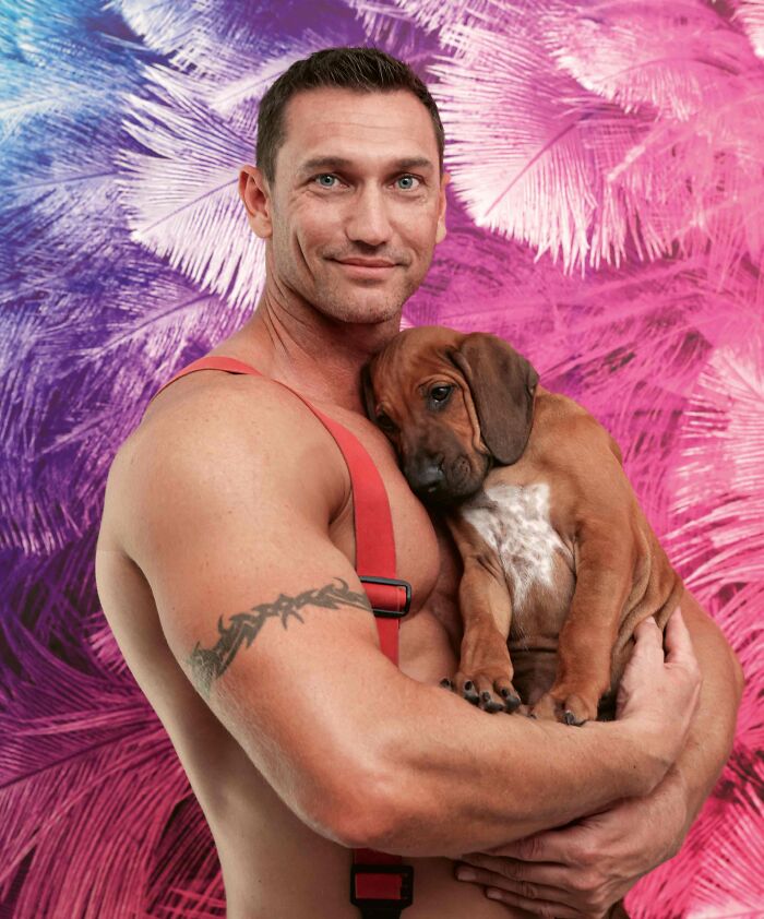 Muscular firefighter holding an adorable puppy against a colorful pink and purple feathered background for Australian calendar.