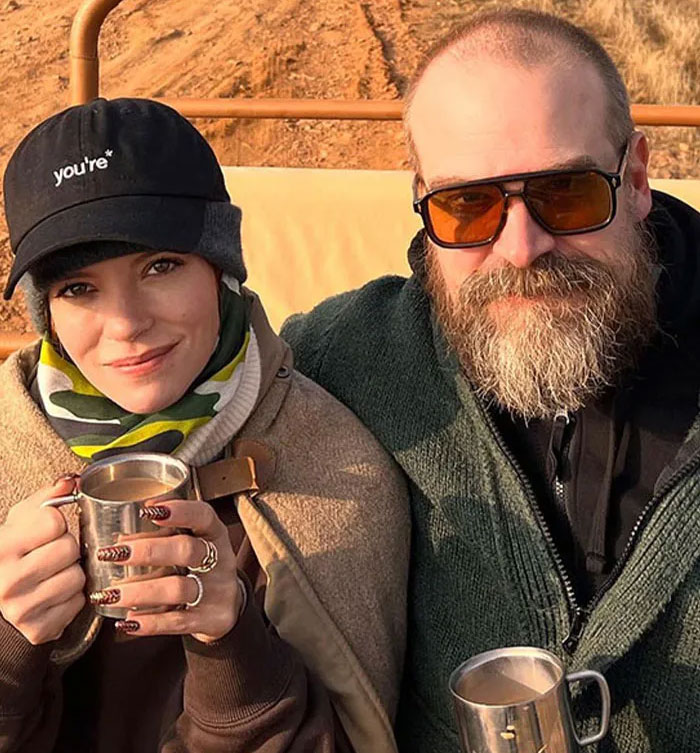 David Harbour and a younger woman sitting outdoors, holding metal mugs during a casual moment.
