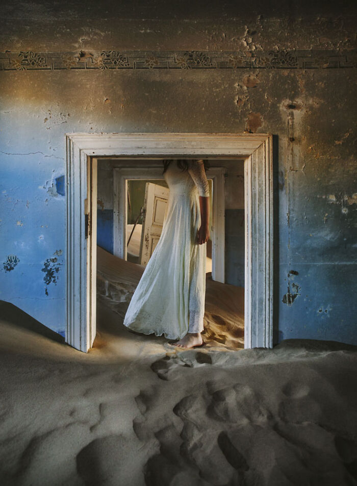 Woman in white dress walking barefoot through desert sand filling abandoned home doorway.