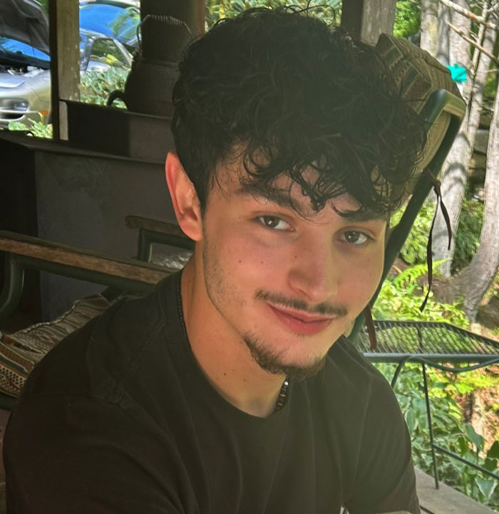Young Florida dad-to-be with curly hair and beard sitting outdoors in a natural setting, smiling softly at the camera. Young Florida dad-to-be with curly hair and beard sitting outdoors in a natural setting, smiling softly at the camera.